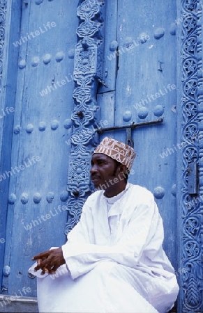  Ein Mann aus Zanzibar in traditionelle Kleidung steht an einer alten Holztuer inmitten der Altstadt Stone Town der Hauptstadt Zanzibar Town auf der Insel Zanzibar welche zu Tansania gehoert.     