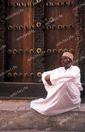  Ein Mann aus Zanzibar in traditionelle Kleidung steht an einer alten Holztuer inmitten der Altstadt Stone Town der Hauptstadt Zanzibar Town auf der Insel Zanzibar welche zu Tansania gehoert.     