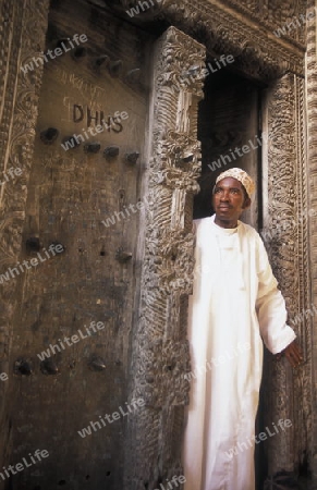  Ein Mann aus Zanzibar in traditionelle Kleidung steht an einer alten Holztuer inmitten der Altstadt Stone Town der Hauptstadt Zanzibar Town auf der Insel Zanzibar welche zu Tansania gehoert.     