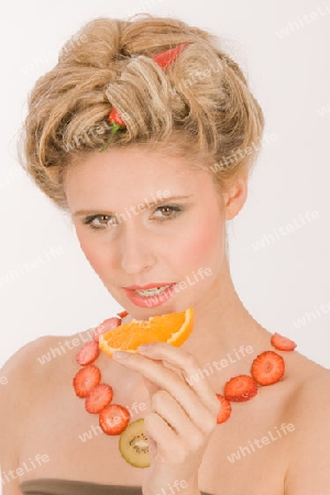 Attraktive blonde junge Frau mit Erdbeere-Kiwi- Kette isst eine Orange / Attraktive blonde junge Frau mit Erdbeere-Kiwi- Kette isst eine Orange