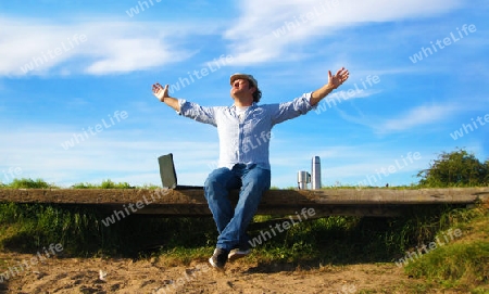 Man sitting on bench with laptop and thermos stretches to the sky - Mann auf Bank sitzend mit Notebook und Thermoskanne streckt sich in Richtung Himmel