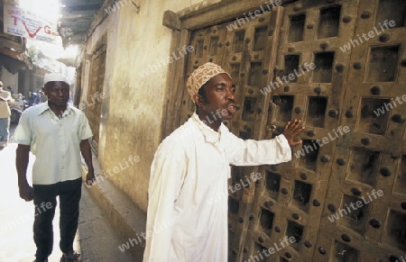  Ein Mann aus Zanzibar in traditionelle Kleidung steht an einer alten Holztuer inmitten der Altstadt Stone Town der Hauptstadt Zanzibar Town auf der Insel Zanzibar welche zu Tansania gehoert.     