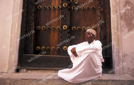  Ein Mann aus Zanzibar in traditionelle Kleidung steht an einer alten Holztuer inmitten der Altstadt Stone Town der Hauptstadt Zanzibar Town auf der Insel Zanzibar welche zu Tansania gehoert.     