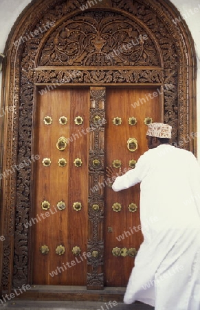  Ein Mann aus Zanzibar in traditionelle Kleidung steht an einer alten Holztuer inmitten der Altstadt Stone Town der Hauptstadt Zanzibar Town auf der Insel Zanzibar welche zu Tansania gehoert.     