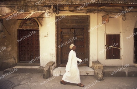  Ein Mann aus Zanzibar in traditionelle Kleidung steht an einer alten Holztuer inmitten der Altstadt Stone Town der Hauptstadt Zanzibar Town auf der Insel Zanzibar welche zu Tansania gehoert.     