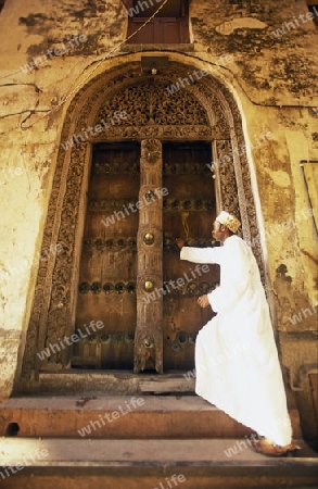  Ein Mann aus Zanzibar in traditionelle Kleidung steht an einer alten Holztuer inmitten der Altstadt Stone Town der Hauptstadt Zanzibar Town auf der Insel Zanzibar welche zu Tansania gehoert.     