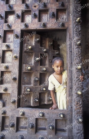  Ein Kind  aus Zanzibar in traditionelle Kleidung steht an einer alten Holztuer inmitten der Altstadt Stone Town der Hauptstadt Zanzibar Town auf der Insel Zanzibar welche zu Tansania gehoert.     