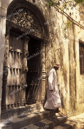  Ein Mann aus Zanzibar in traditionelle Kleidung steht an einer alten Holztuer inmitten der Altstadt Stone Town der Hauptstadt Zanzibar Town auf der Insel Zanzibar welche zu Tansania gehoert.     